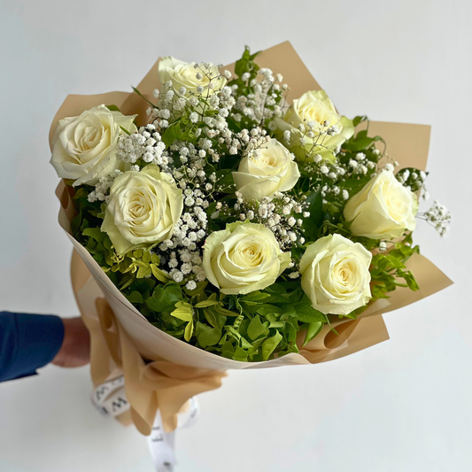 A bouquet of white roses and baby's breath with a generous amount of foliage, wrapped in brown paper for Same-Day Delivery in Karachi